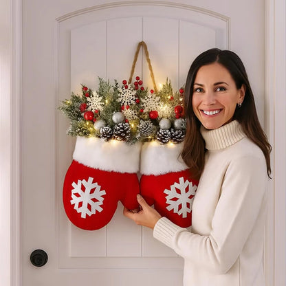Smiling woman holds glove-shaped Christmas garland with red mitten accents, white cuffs, greenery, pinecones, and lights on a door.